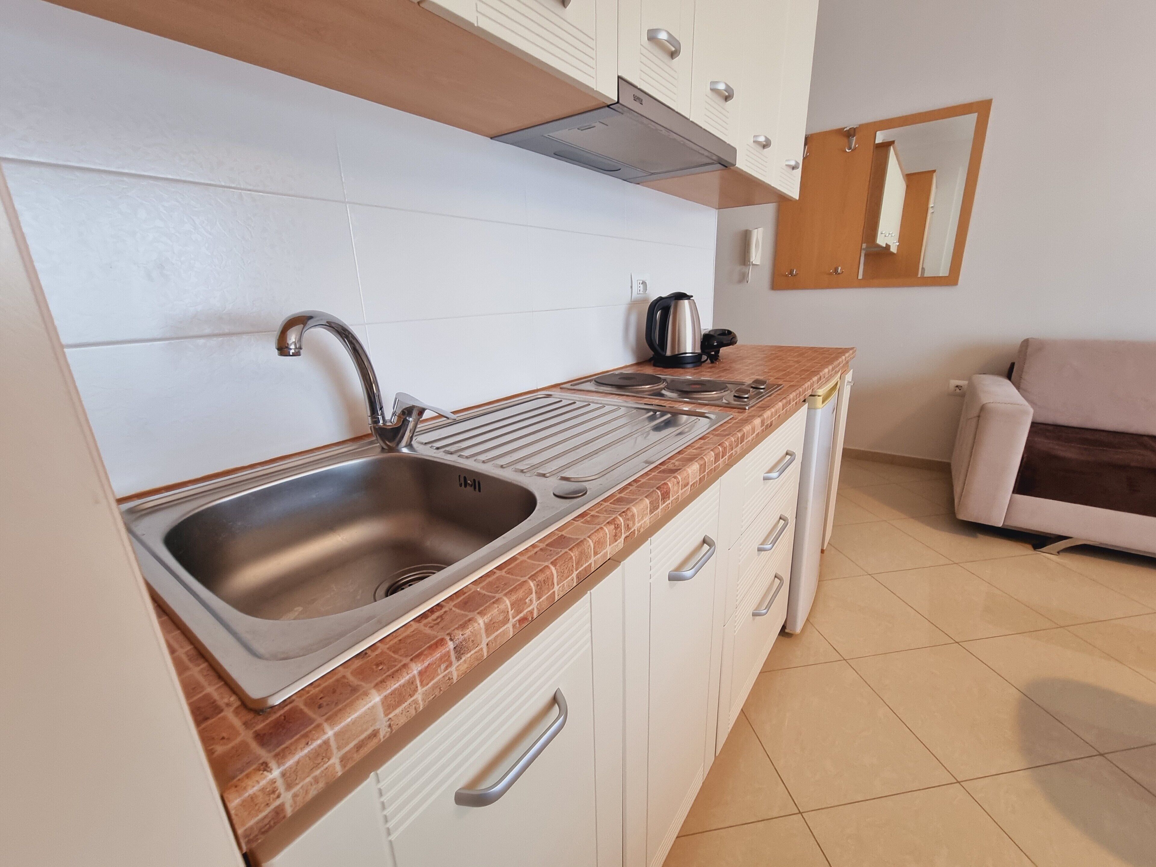 Una cocina con un lavamanos de acero inoxidable, una tetera y una estufa. Hay un sofá color crema y un espejo en la pared.