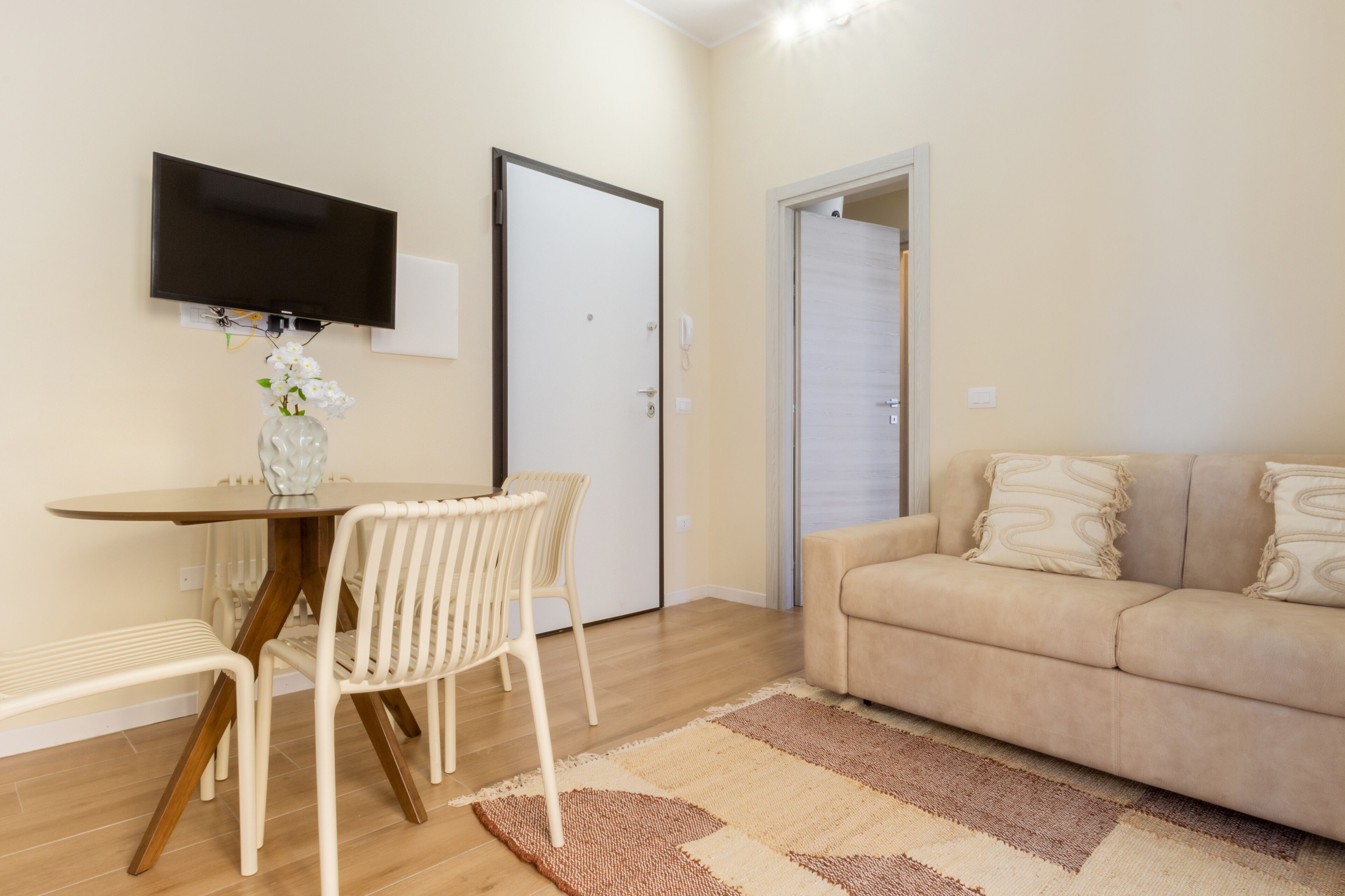 A living room with a beige sofa, a dining table with chairs, a flat-screen TV mounted on the wall, and a door leading to another room.