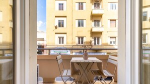 Een balkon met een tafel en stoelen, een vaas met bloemen en uitzicht op een geel gebouw met meerdere ramen.