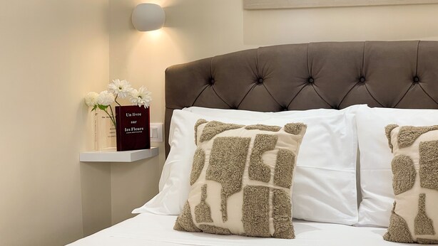 Een net opgemaakt bed met witte lakens en twee decoratieve kussens met een getextureerd ontwerp. Een nachtkastje bevat een boek en een vaas met witte bloemen.