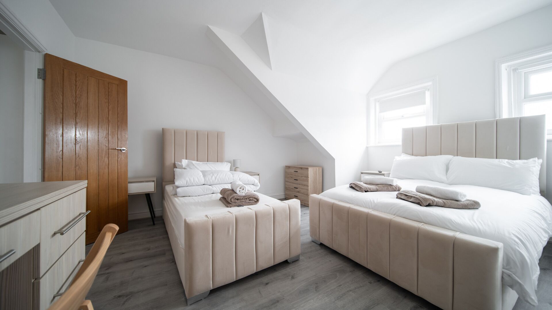 Une chambre moderne avec un plafond en pente, deux lits avec des têtes de lit assorties, une porte en bois et une petite table de chevet.