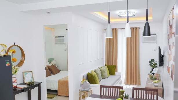 A modern hotel room with a dining area, a bed with green and brown pillows, and a small table with a plant.
