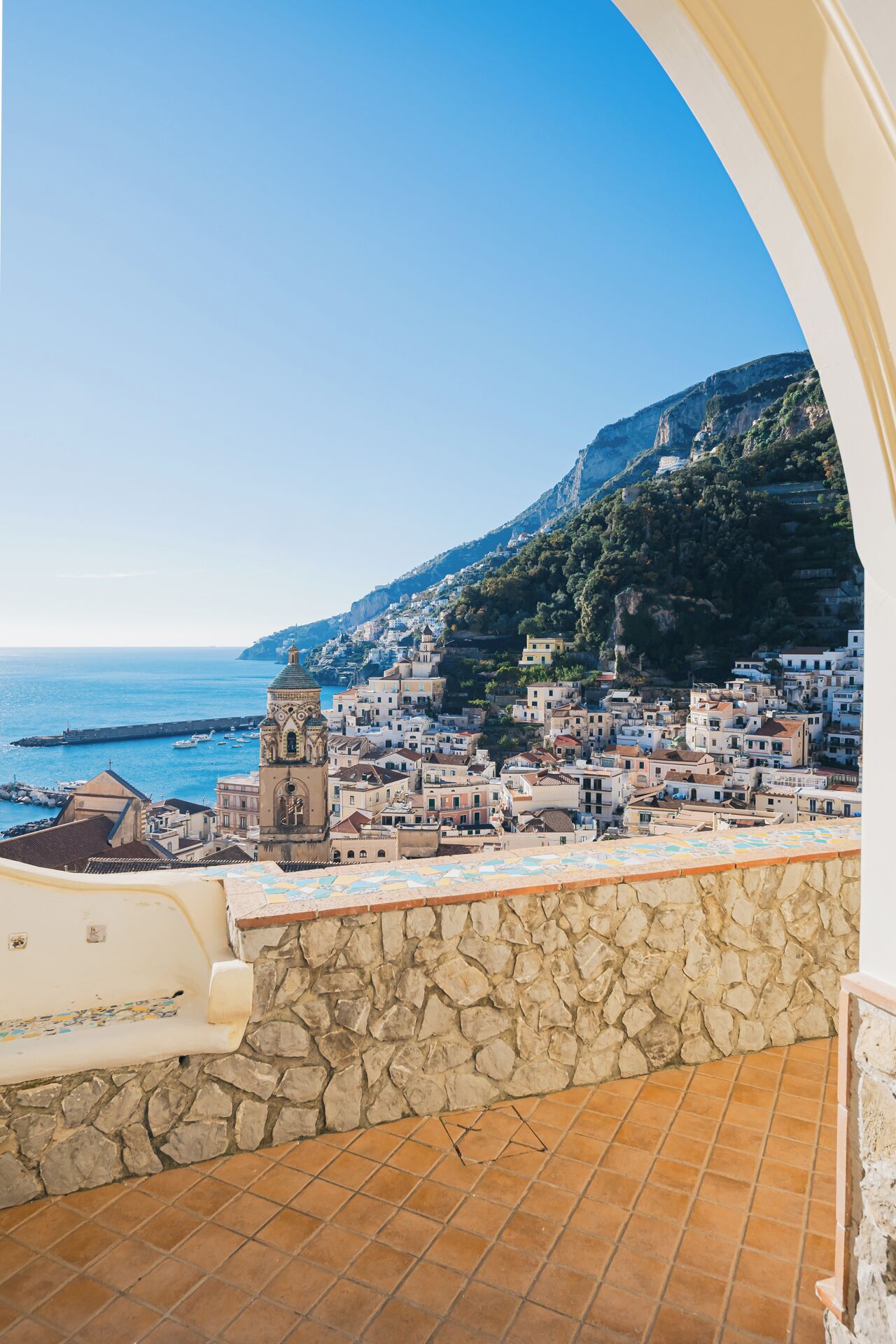 A coastal town with a prominent bell tower, terracotta-tiled balconies, and a stone wall.