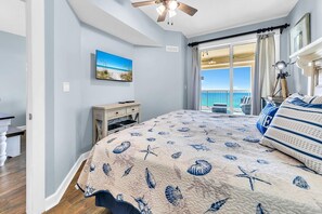 A bedroom with a large bed, a desk, a chair, a ceiling fan, a window with a view of the ocean, and a painting on the wall. - Grand Panama 1-1807 (Panama City Beach)