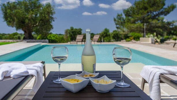 A poolside table with two glasses of white wine, a bottle of white wine, and bowls of chips.