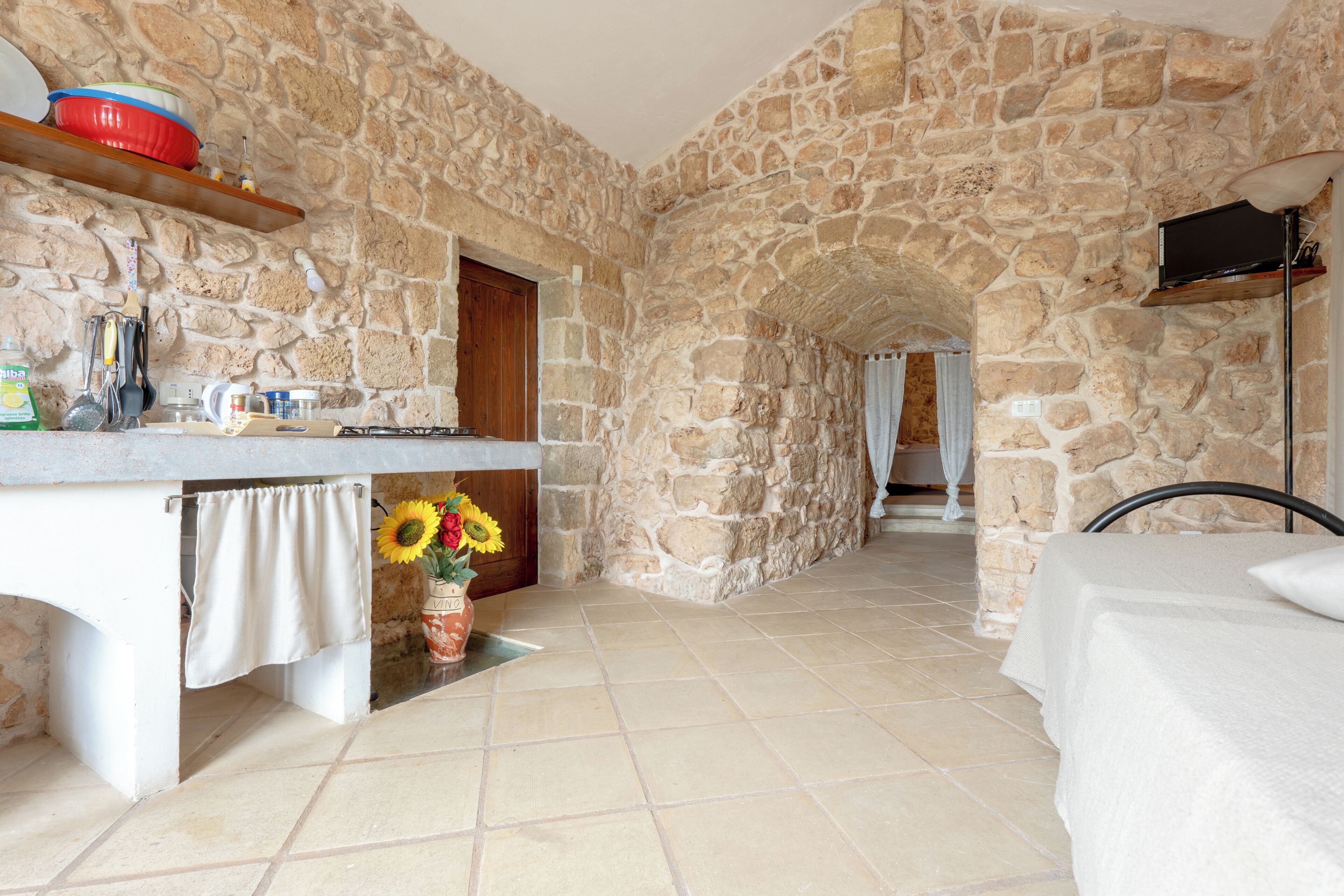 A stone-walled room with a kitchen area, a bed, and a hallway leading to another room.