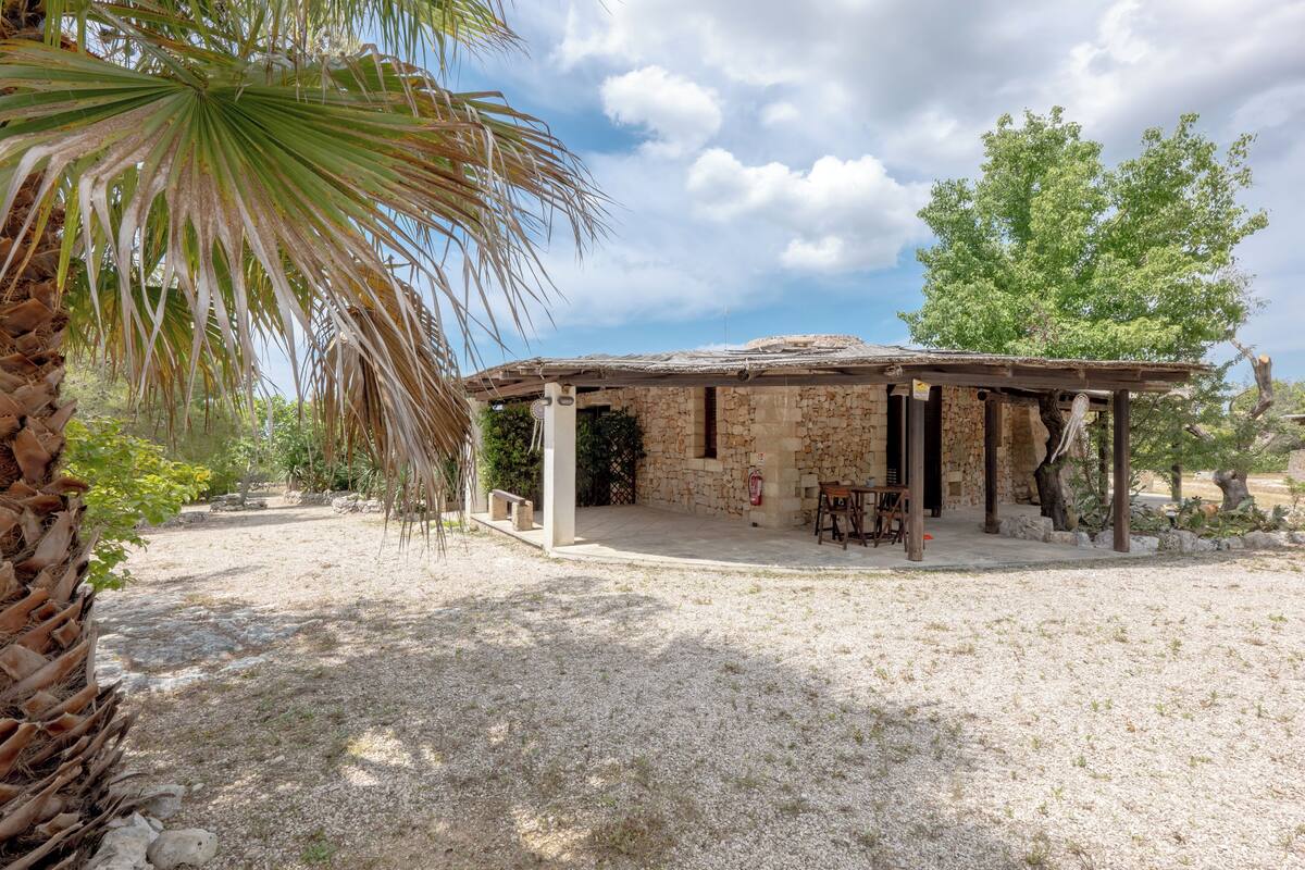 Un bâtiment rustique en pierre, doté d’un toit de chaume, d’un espace extérieur abrité avec des sièges et d’un palmier au premier plan.