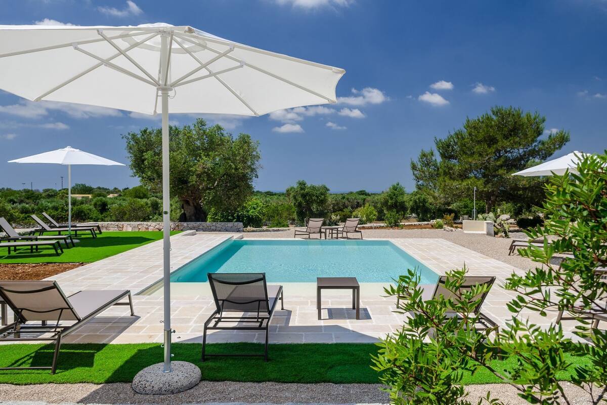 Une piscine entourée de verdure, avec des chaises longues et des parasols, sous un ciel dégagé.