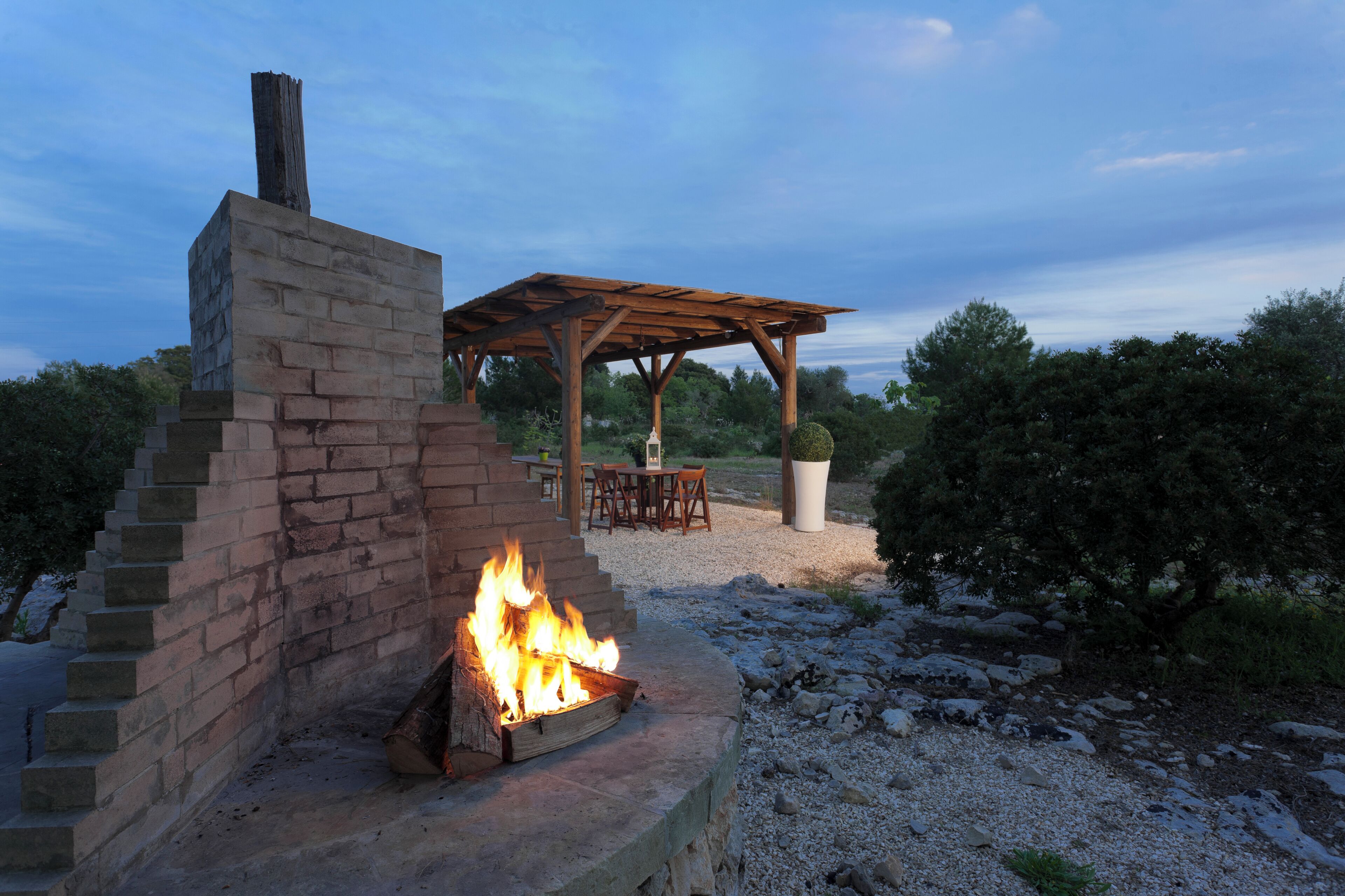 A brick fire pit with flames, a wooden pergola, and a table and chairs set outdoors.