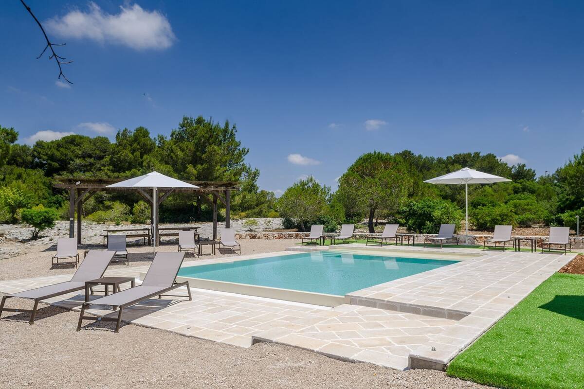 Une piscine rectangulaire entourée d’une terrasse en pierre et de verdure, avec des chaises longues et des parasols.