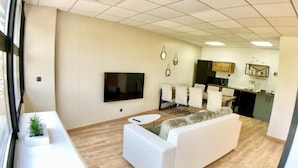 A modern living room with a white sofa, a small round table, and a flat-screen TV mounted on the wall. There is a kitchen area with a dining table and chairs visible in the background. - New house Ovalie Montpellier near Stade de Rugby (Montpellier)