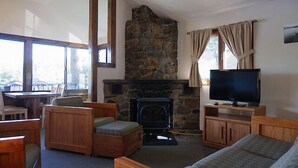 A cozy living room with a stone fireplace, wooden furniture, a television, and large windows. - Come stay at our four season resort! (Weare)