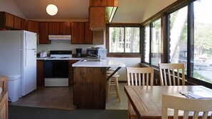 A compact kitchen with wooden cabinets, a white refrigerator, a stove, and a microwave. There is a wooden island with a sink and chairs around it. Large windows provide natural light and a view of the outdoors. - Come stay at our four season resort! (Weare)
