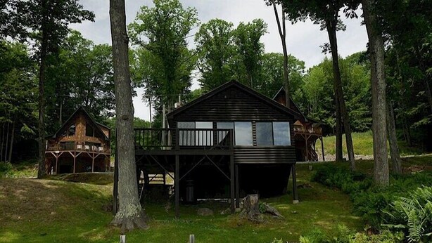 A rustic cabin with a wooden deck and a covered porch, surrounded by trees. - Come stay at our four season resort! (Weare)