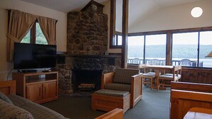A cozy living room with a stone fireplace, wooden furniture, a flat-screen TV, and large windows offering a view of the outdoors. - Come stay at our four season resort! (Weare)