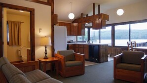 A cozy living room with a sofa, two armchairs, a small table with a lamp, wooden cabinets, and a kitchen area with a view of the outdoors. - Come stay at our four season resort! (Weare)