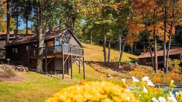 A rustic cabin with a wooden deck and a small porch, surrounded by trees with autumn foliage. - Come stay at our four season resort! (Weare)