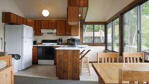 A compact kitchen with wooden cabinets, a white refrigerator, a stove, and a dining area with wooden chairs. - Come stay at our four season resort! (Weare)