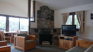 A cozy living room with a stone fireplace, wooden furniture, a television, and large windows. - Come stay at our four season resort! (Weare)