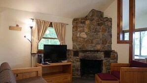 A cozy living room with a stone fireplace, a television on a wooden stand, and a window with curtains. - Come stay at our four season resort! (Weare)