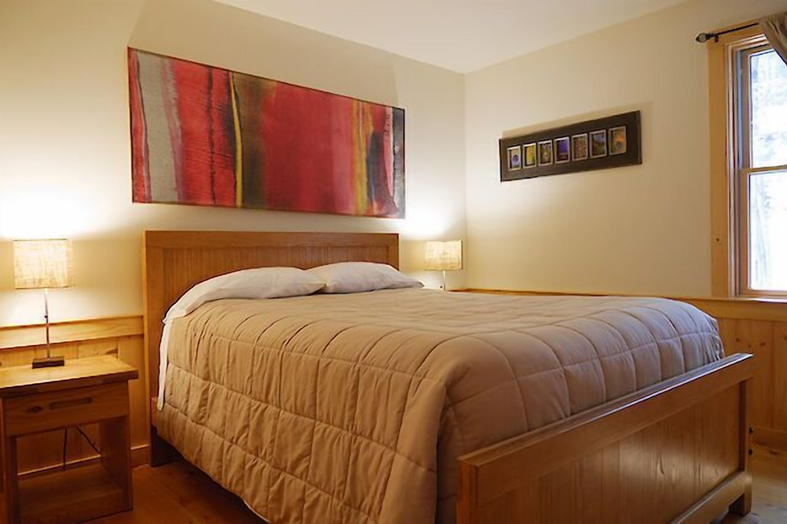 A bedroom with a wooden bed, two bedside tables, a large abstract painting, and a framed photo collection on the wall.