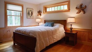 A bedroom with a wooden bed, two bedside lamps, a window with blinds, and wall art. - Come stay at our four season resort! (Weare)