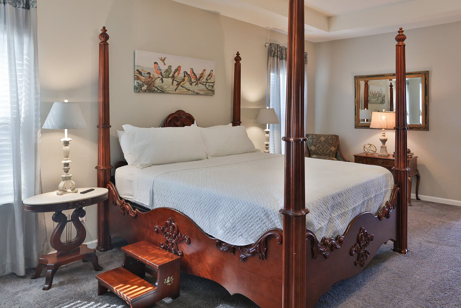 A four-poster bed with a white quilt, a nightstand with a lamp, a chair, and a painting of birds on the wall.