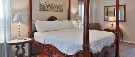 A four-poster bed with a white quilt, a nightstand with a lamp, a chair, and a painting of birds on the wall.