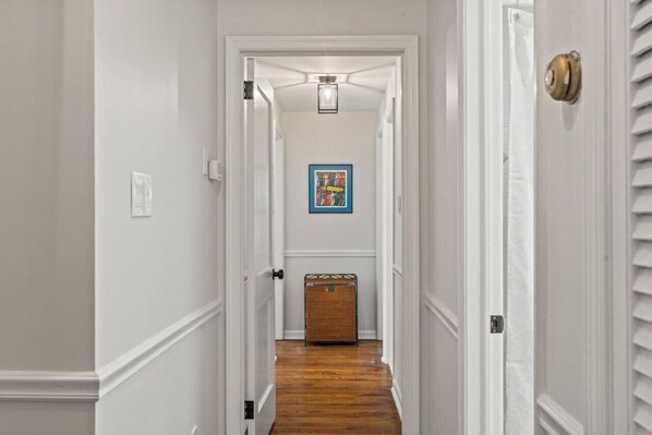 A narrow hallway with white walls, a wooden floor, a ceiling light, and a framed picture on the wall.
