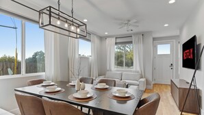 Une salle à manger moderne avec une grande table dressée pour six personnes, entourée de sièges confortables. On peut voir un ventilateur de plafond et des spots encastrés.
