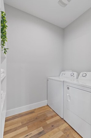 A laundry room with two white washing machines, wooden flooring, and a green plant on the left wall. - Modern 3BR Home | 5 Min to Downtown & SXSW (Austin)