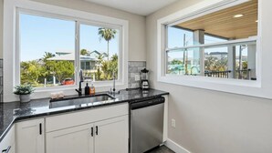 A kitchen with a dishwasher, a blender, and a window with a view of houses and palm trees. - Entire 6BR Beach Duplex | Walk to Beach | Sleeps 12 (Bradenton Beach)