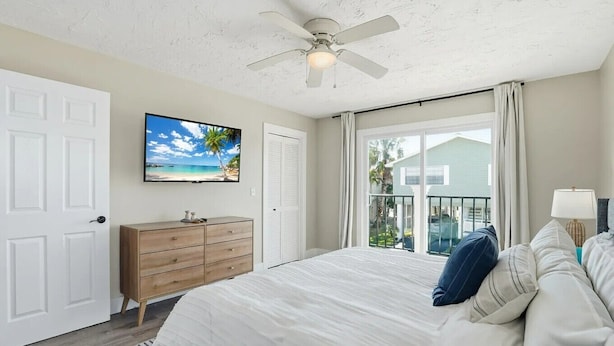 A bedroom with a bed, a dresser, a TV, a ceiling fan, and a view of a house through a window. - Entire 6BR Beach Duplex | Walk to Beach | Sleeps 12 (Bradenton Beach)