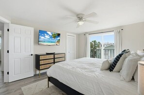 A bedroom with a bed, a television on the wall, a ceiling fan, and a window with a view.