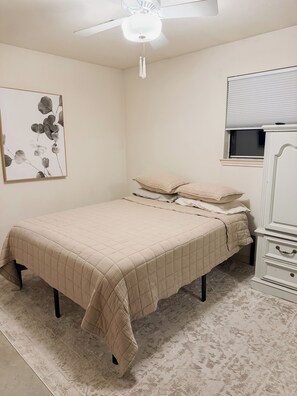 Quarto com cama, ventoinha no teto, janela com persianas, mesa de cabeceira e uma obra de arte na parede.