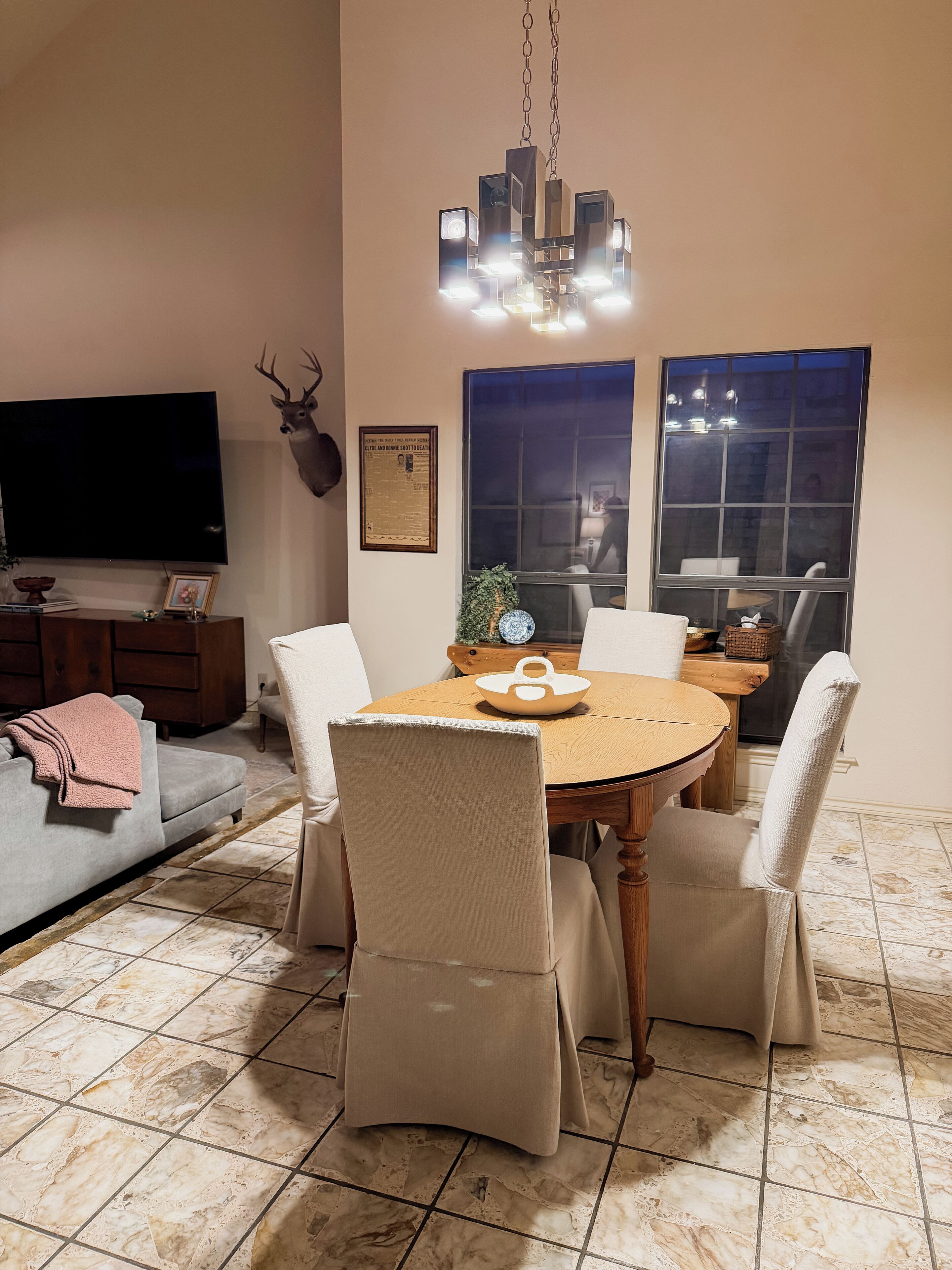 Un comedor con una mesa redonda de madera, cuatro sillas tapizadas, una araña, una cabeza de venado montada, un televisor y un certificado enmarcado en la pared.