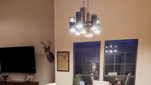 A dining area with a round wooden table, four upholstered chairs, a chandelier, a mounted deer head, a television, and a framed certificate on the wall.