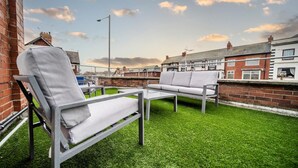 A rooftop terrace with a metal table and two white cushioned chairs, surrounded by a green lawn and brick walls.