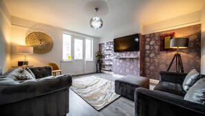 A modern living room with a grey sofa set, a patterned rug, a flat-screen TV mounted on the wall, and a floor lamp.
