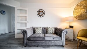 A modern living room with a grey sofa, patterned cushions, a decorative wall clock, a lamp, and a chair.