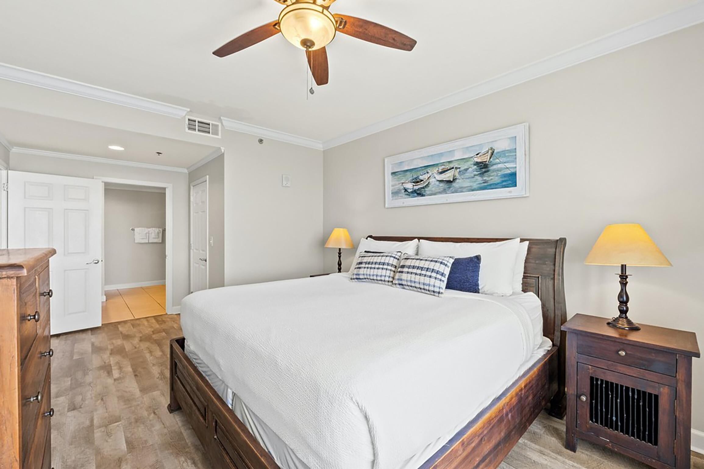 A bedroom with a large bed, two bedside lamps, a ceiling fan, and a painting on the wall.