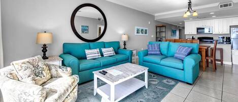 Una sala de estar con sofás color turquesa, una mesa de centro blanca y un sillón estampado. Se ve una cocina al fondo.