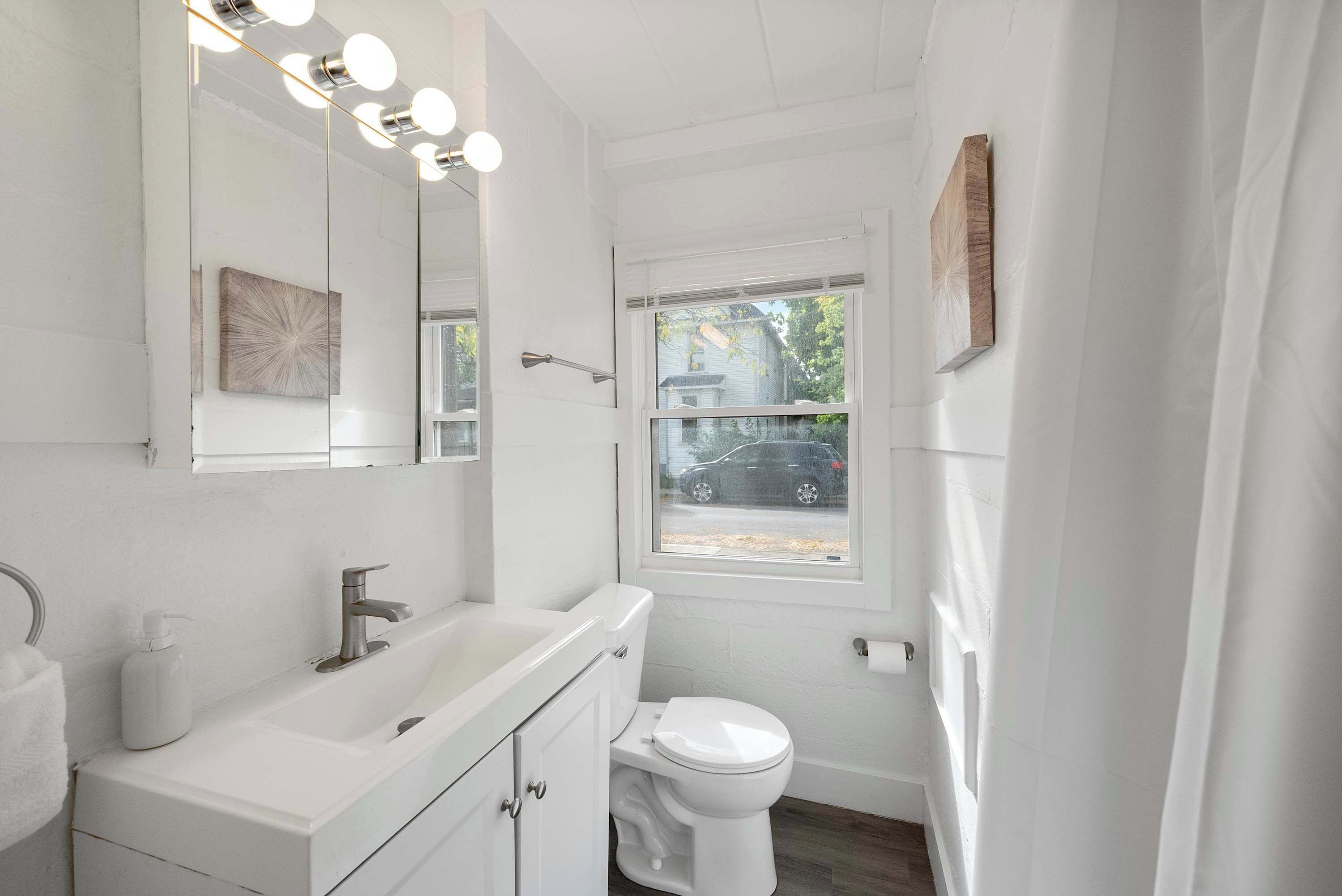 A bathroom with a white vanity, a large mirror with lights, a toilet, and a window with a view of the outside.