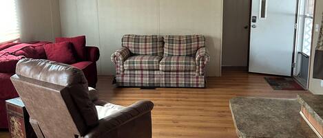A spacious living room with a ceiling fan, a red sofa, a plaid sofa, a recliner, and a kitchen area with a granite countertop.