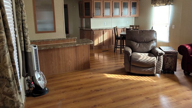A spacious living room with a wooden floor, a leather armchair, a dining table with chairs, and a kitchen area with wooden cabinets and a countertop.