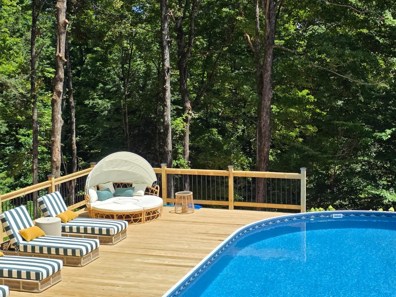 Zona junto a la piscina con sillas de descanso y una tumbona, rodeada de altos árboles.