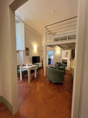A spacious living area with a dining table, green armchairs, a TV, and a kitchen in the background.