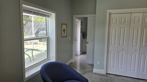A room with a large window, a chair, a door, and a hallway leading to a bathroom.