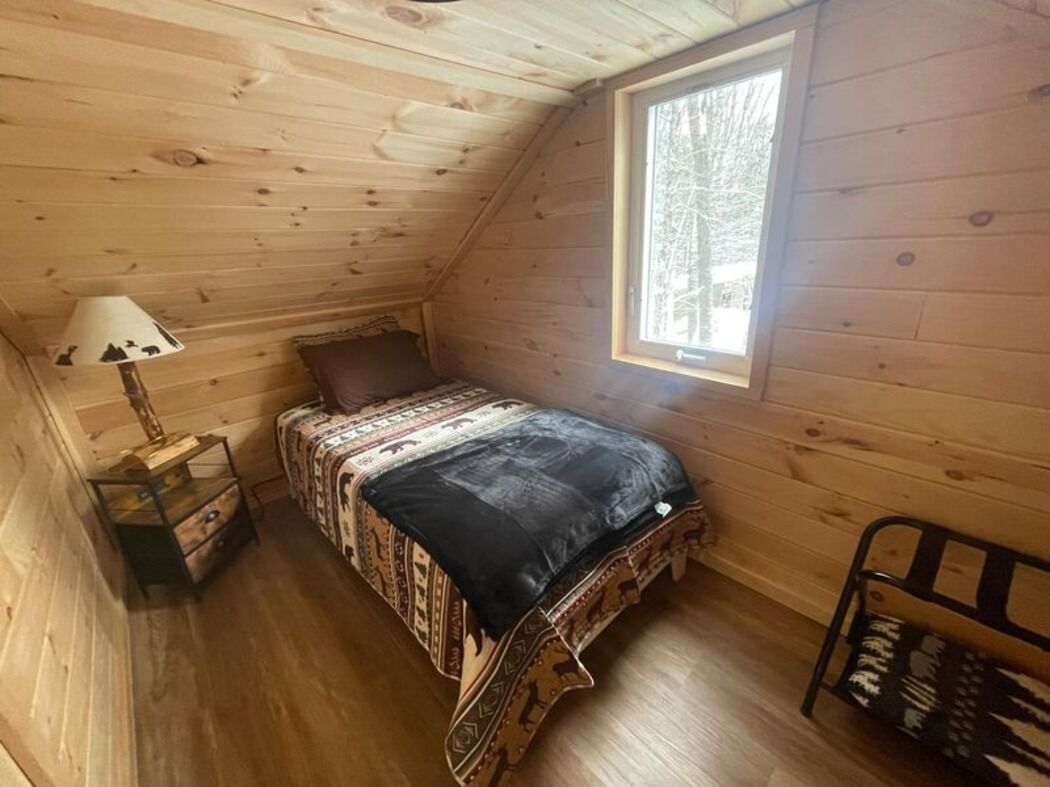 Una habitación pequeña y acogedora, con techo inclinado, una cama individual con ropa de cama estampada, una mesita de noche con lámpara y una ventana con vistas a los árboles.