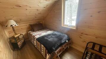Una habitación pequeña y acogedora, con techo inclinado, una cama individual con ropa de cama estampada, una mesita de noche con lámpara y una ventana con vistas a los árboles.